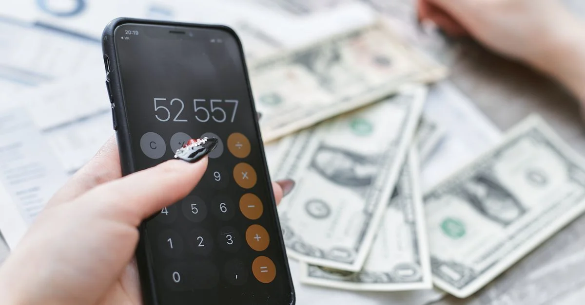 Gros plan sur des mains utilisant la calculatrice d'un smartphone à côté de billets de dollar sur une table. Concept de planification financière.