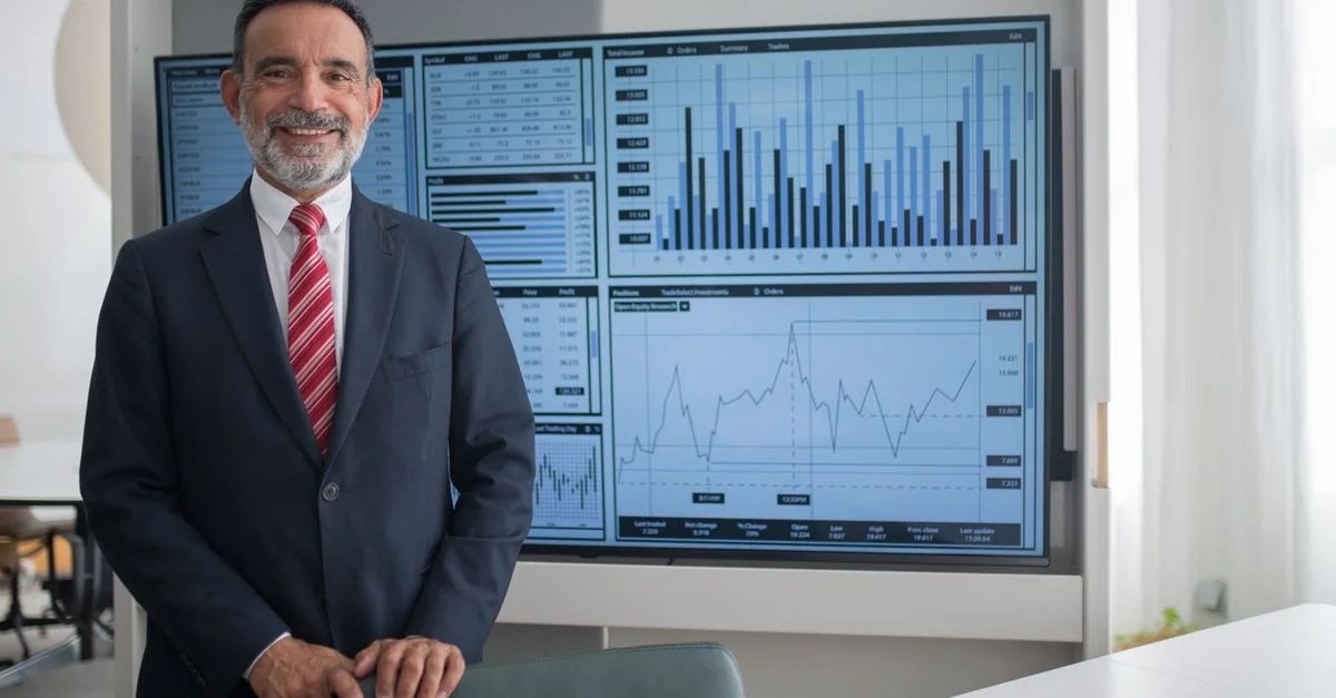 Homme d'affaires souriant dans un bureau analysant des graphiques financiers sur un grand écran, démontrant le succès professionnel.