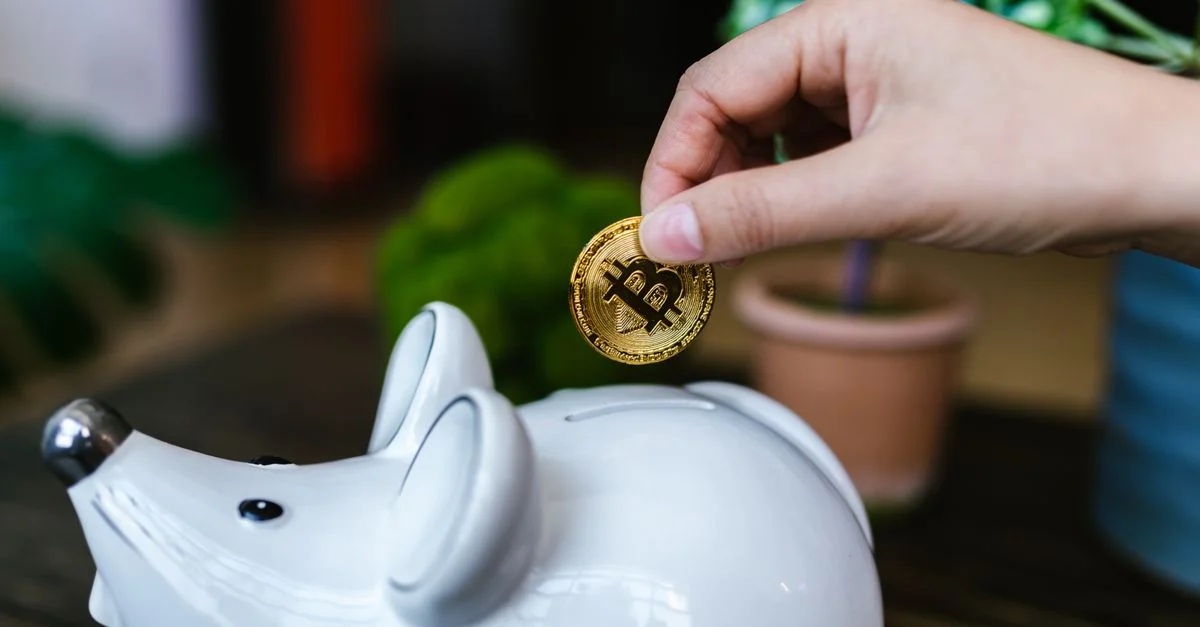 Une main plaçant une pièce de Bitcoin dans une tirelire blanche symbolisant l'épargne.
